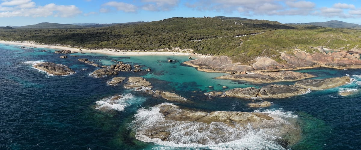 A coastal landscape with a sandy beach, rocky outcrops, and a green hill in the distance.