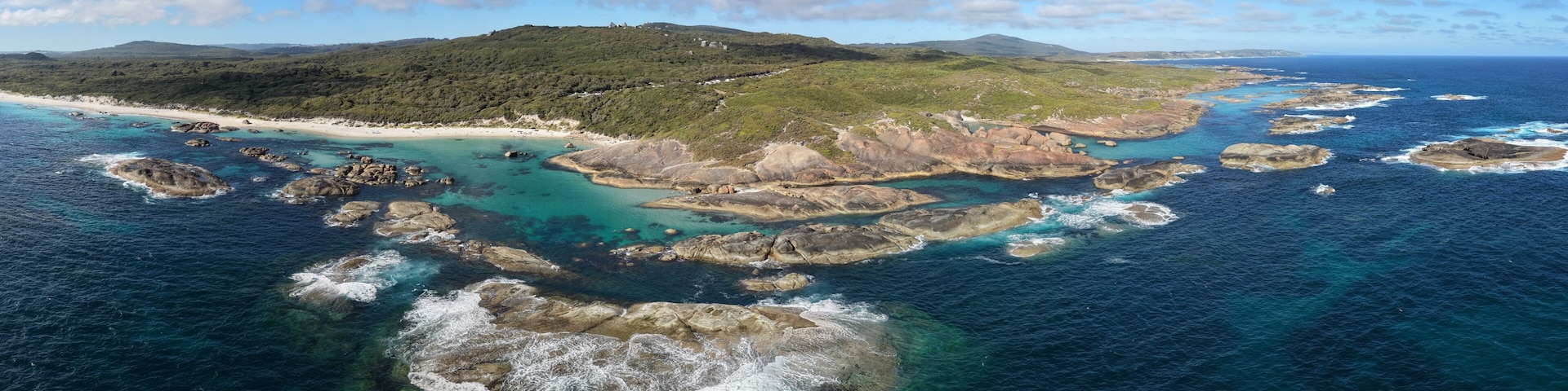 A coastal landscape with a sandy beach, rocky outcrops, and a green hill in the distance.