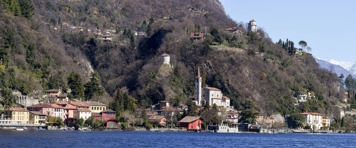 A lakeside village with buildings on a hillside, a lighthouse, and snow-capped mountains in the background.