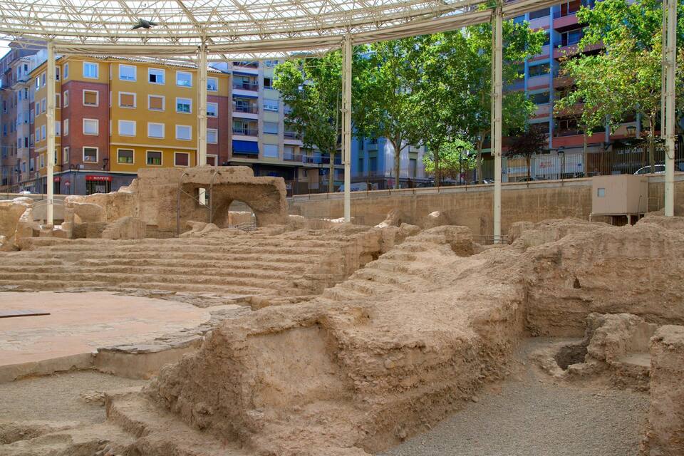 Ruínas antigas com degraus de pedra e um grande arco, rodeadas por edifícios modernos e árvores.