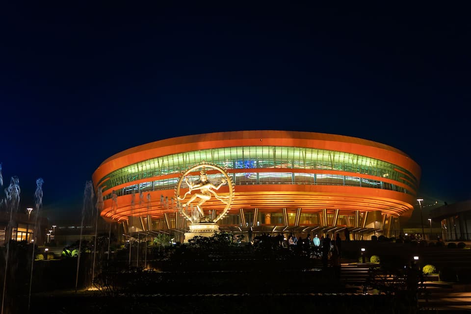 Een groot, rond stadion met een opvallend logo in het midden, omgeven door waterpartijen en verlicht met kunstmatige verlichting.