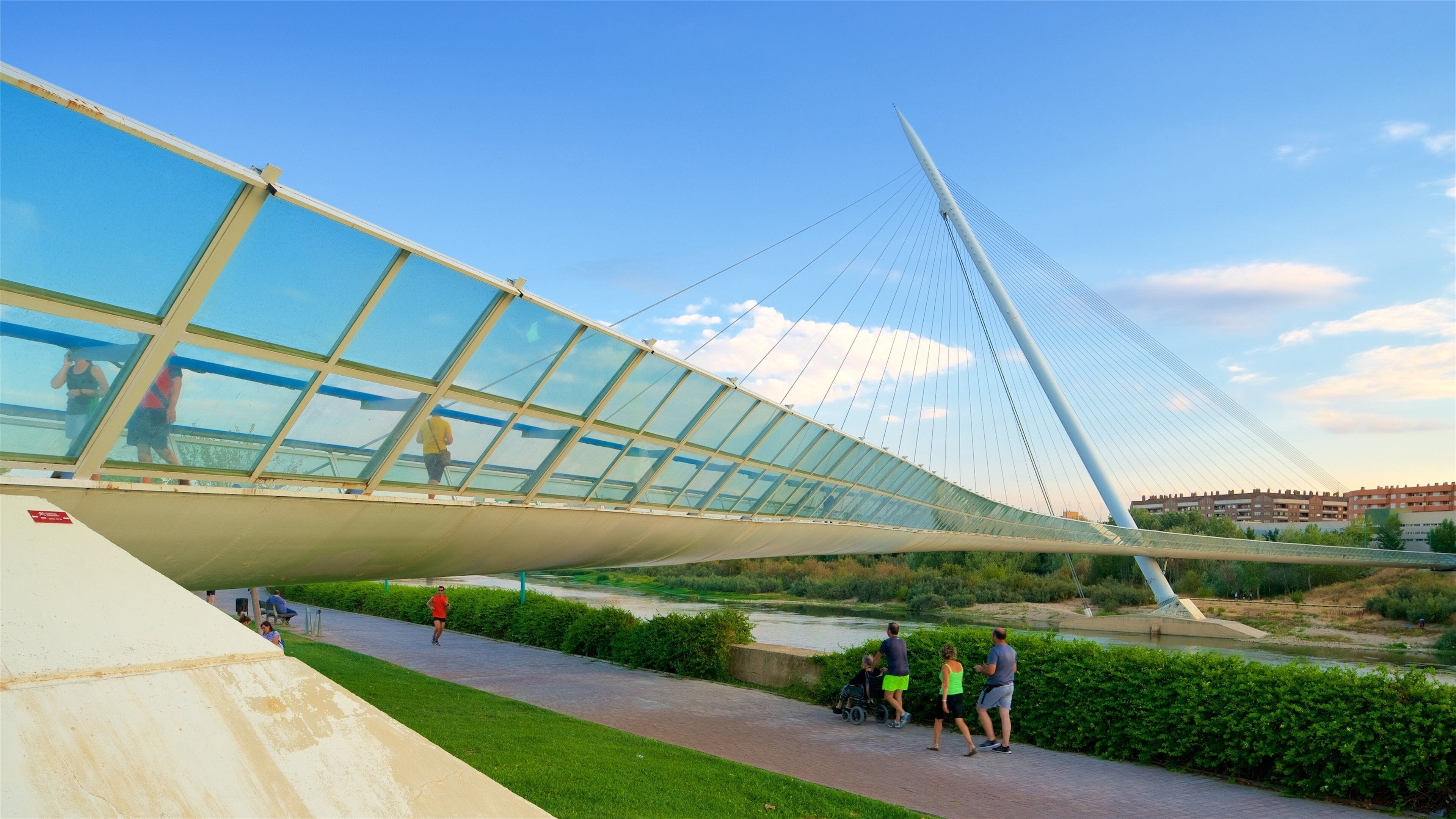 A modern pedestrian bridge with a glass canopy, overlooking a river and greenery.
