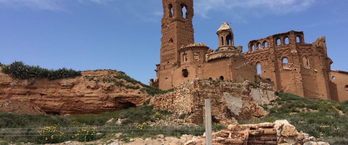 A large, ancient stone structure with a tall tower and multiple arches, surrounded by rocky terrain and sparse vegetation.
