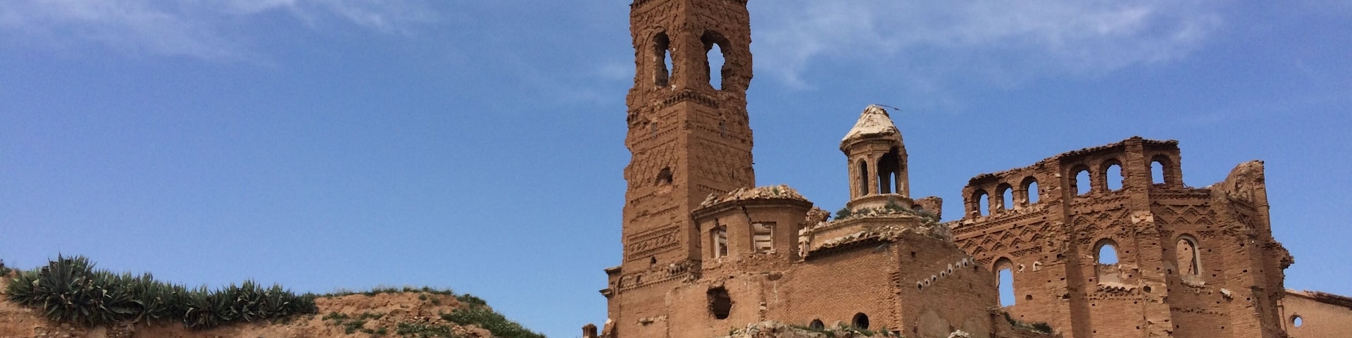 A large, ancient stone structure with a tall tower and multiple arches, surrounded by rocky terrain and sparse vegetation.