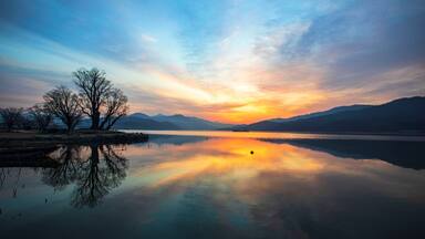 A calm lake with a sunset, trees, and distant mountains.