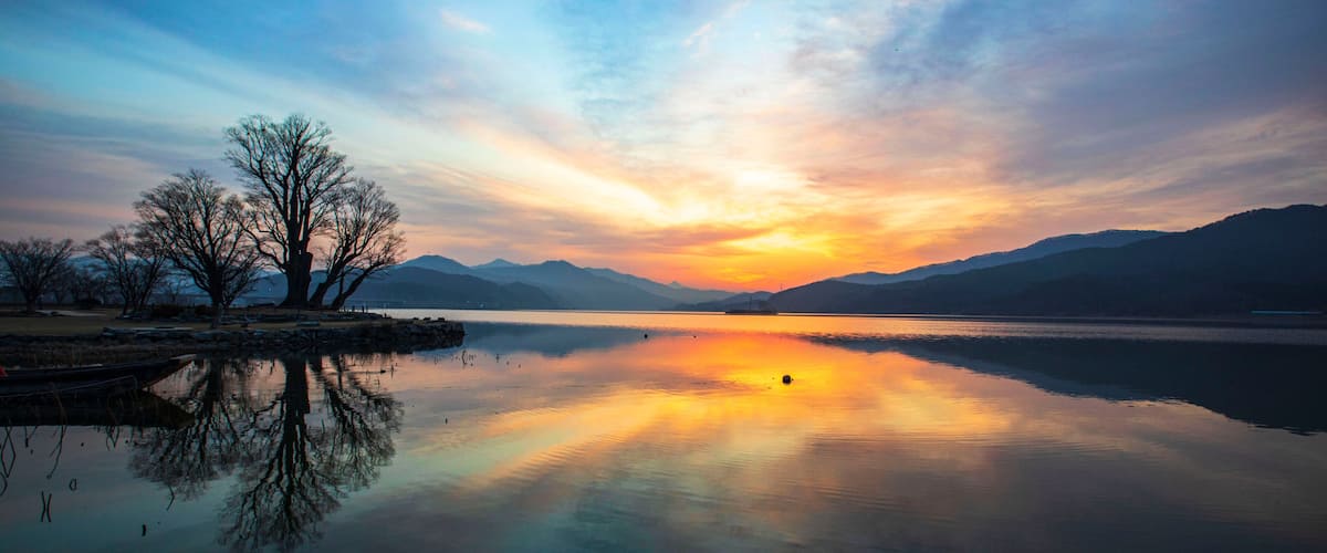 A calm lake with a sunset, trees, and distant mountains.