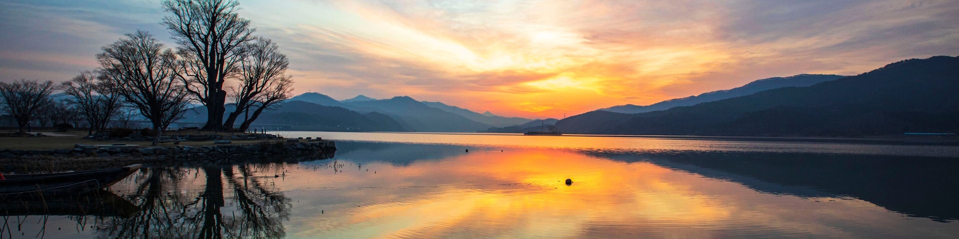 A calm lake with a sunset, trees, and distant mountains.
