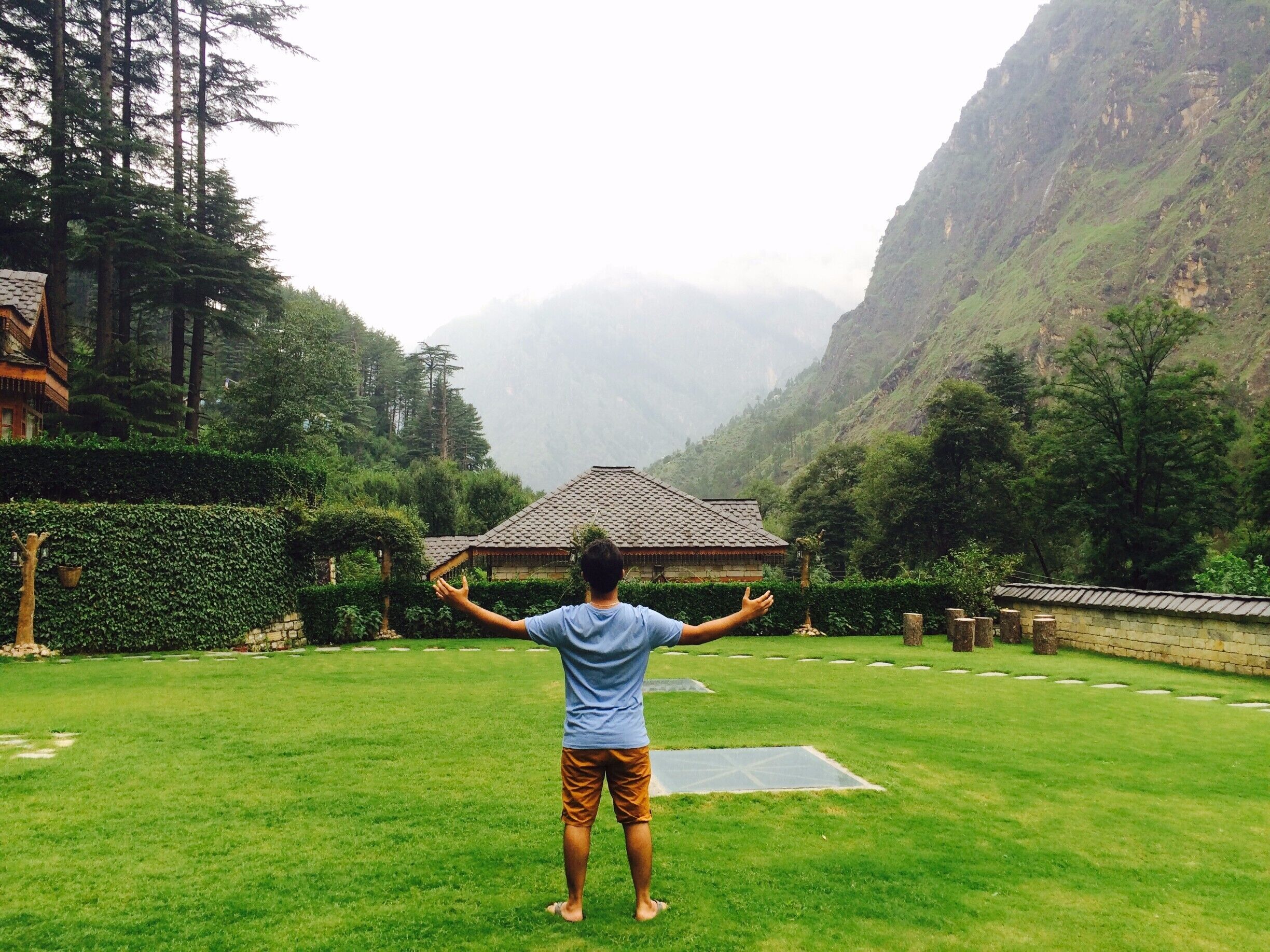 A person standing in a grassy area with a traditional building in the background and mountains in the distance.