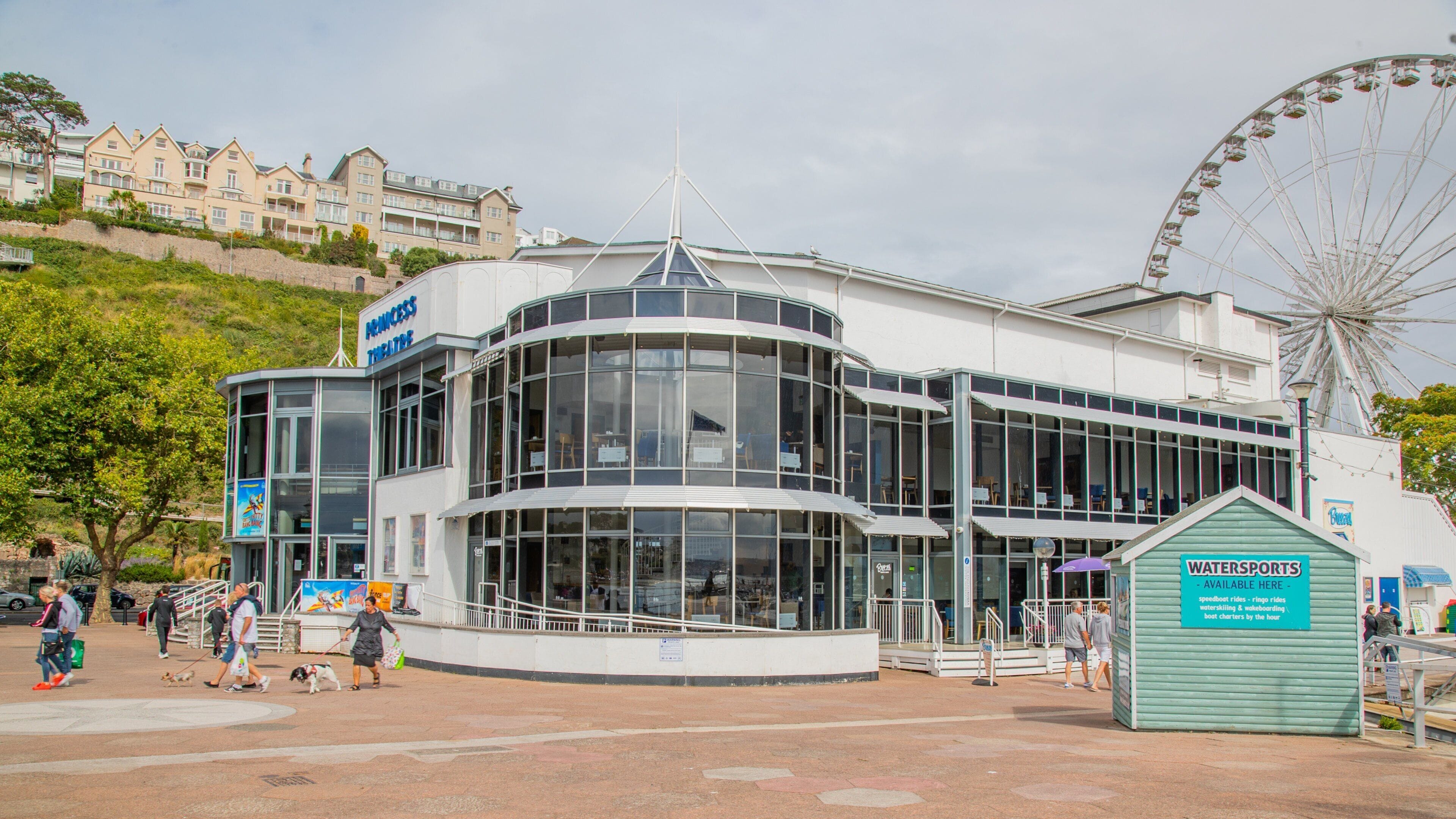 Un edificio moderno con un cartel que dice 'Princess Theatre' y un puesto cercano de 'Watersports' que anuncia actividades como paseos en lancha rápida y wakeboard.