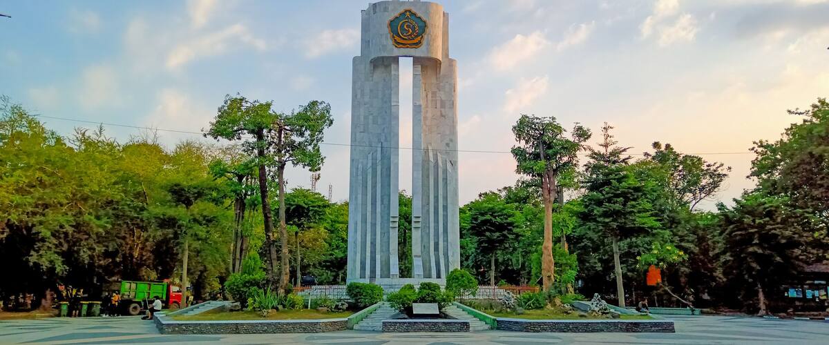 A tall monument with a circular emblem at the top, surrounded by a landscaped area with trees and a paved walkway.