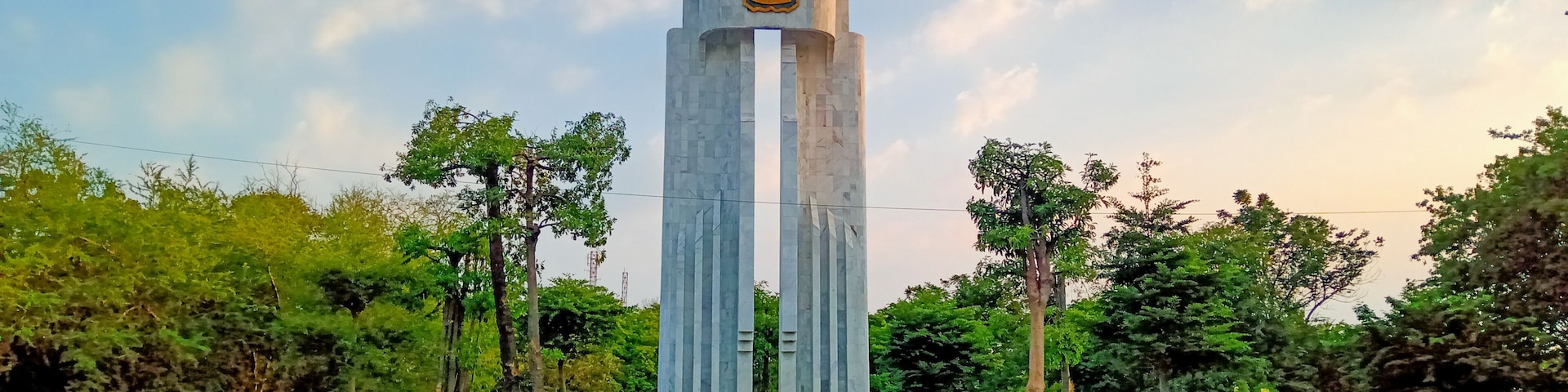 A tall monument with a circular emblem at the top, surrounded by a landscaped area with trees and a paved walkway.