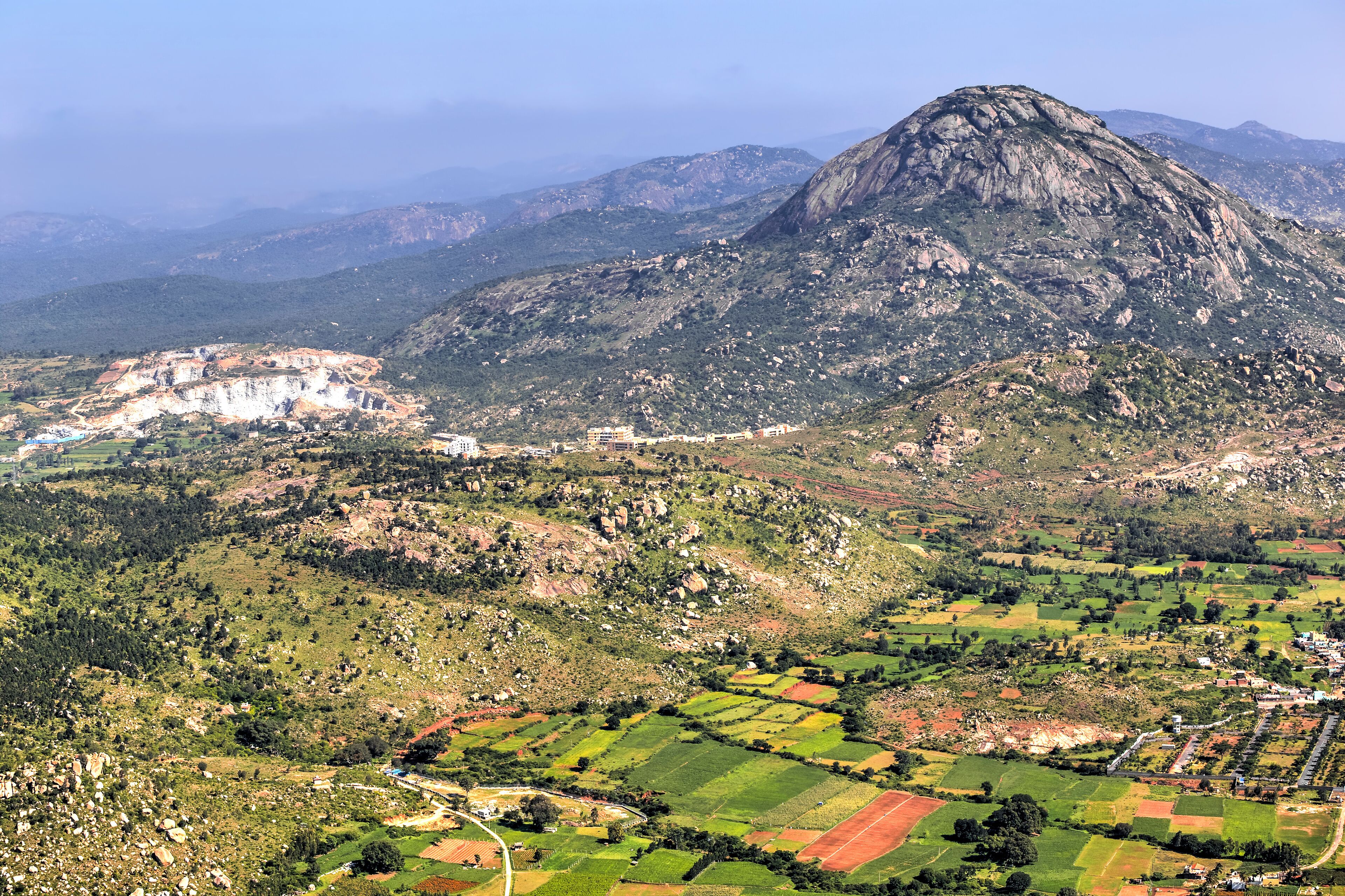 A mountainous landscape with terraced fields, a prominent rocky peak, and a small settlement with buildings.