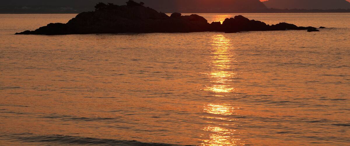 A sunset over a calm sea with a rocky outcrop in the foreground and mountains in the background.
