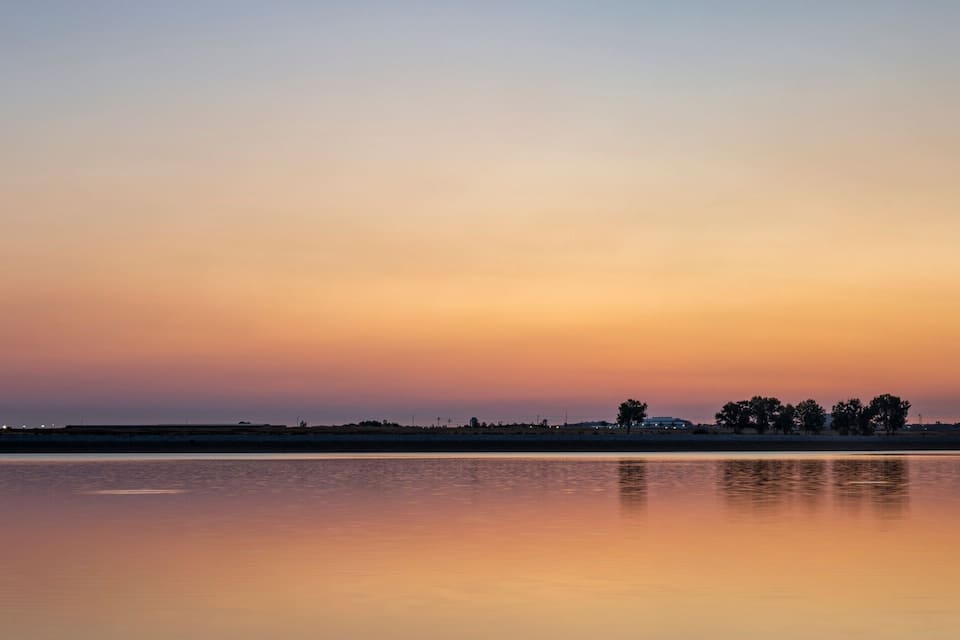 Um lago tranquilo ao pôr do sol, com linha do horizonte e silhuetas de árvores e edifícios.