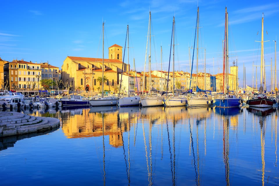 A marina with multiple sailboats docked, reflecting in calm water, surrounded by historic buildings.