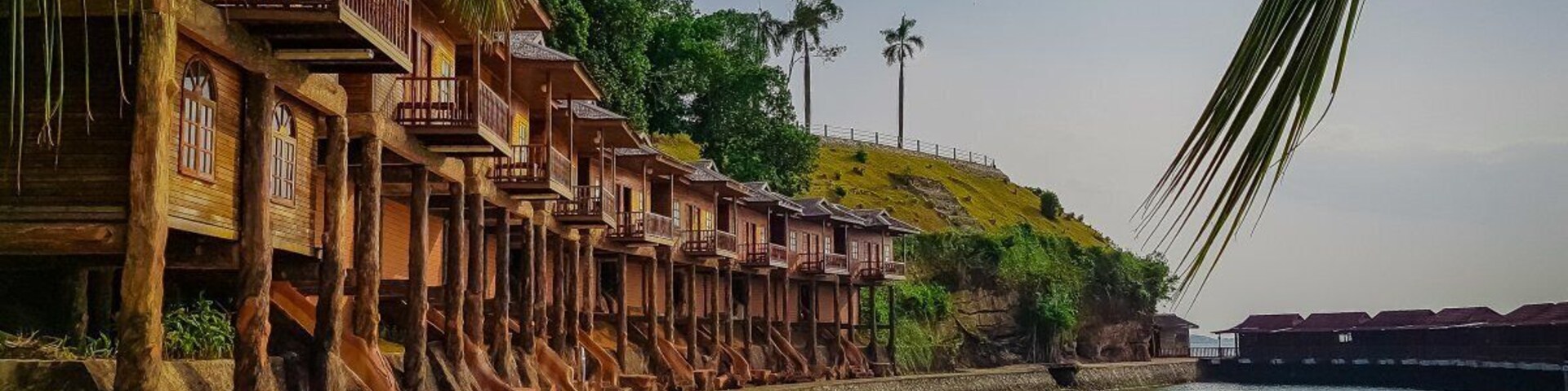 Un paseo marítimo con casas de madera sostenidas por pilotes, una playa de arena y un río tranquilo.