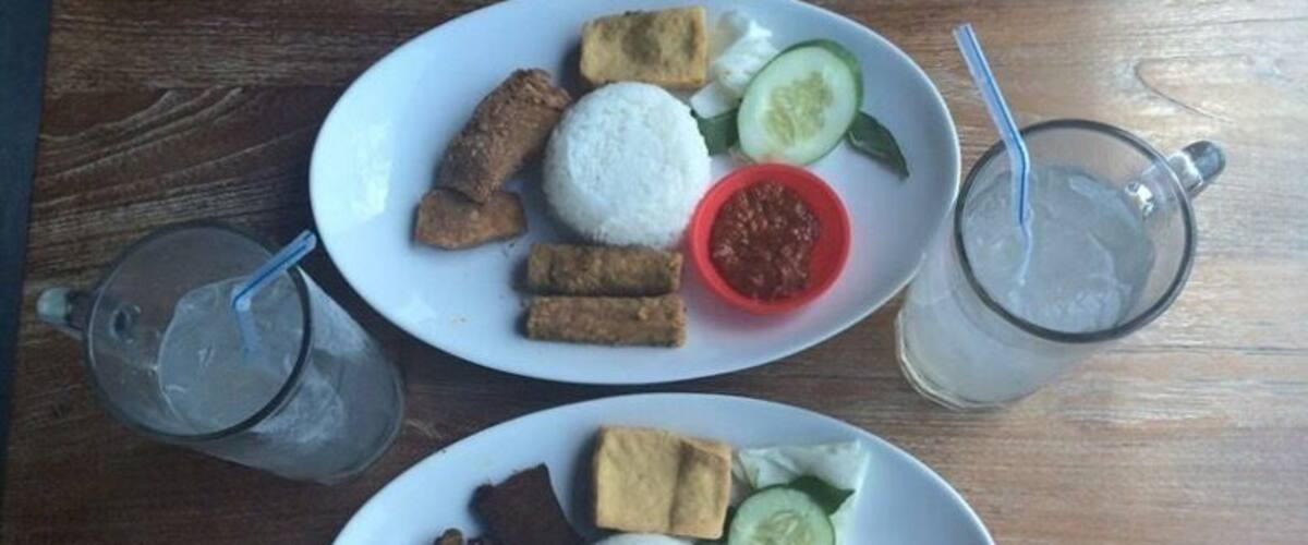 Two plates of food with rice, vegetables, and meat, and two glasses of iced drink on a wooden table.