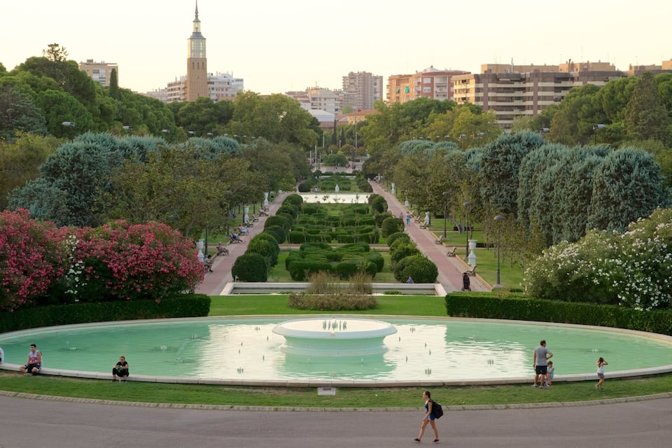 A park with a fountain, trees, and a distant tower.