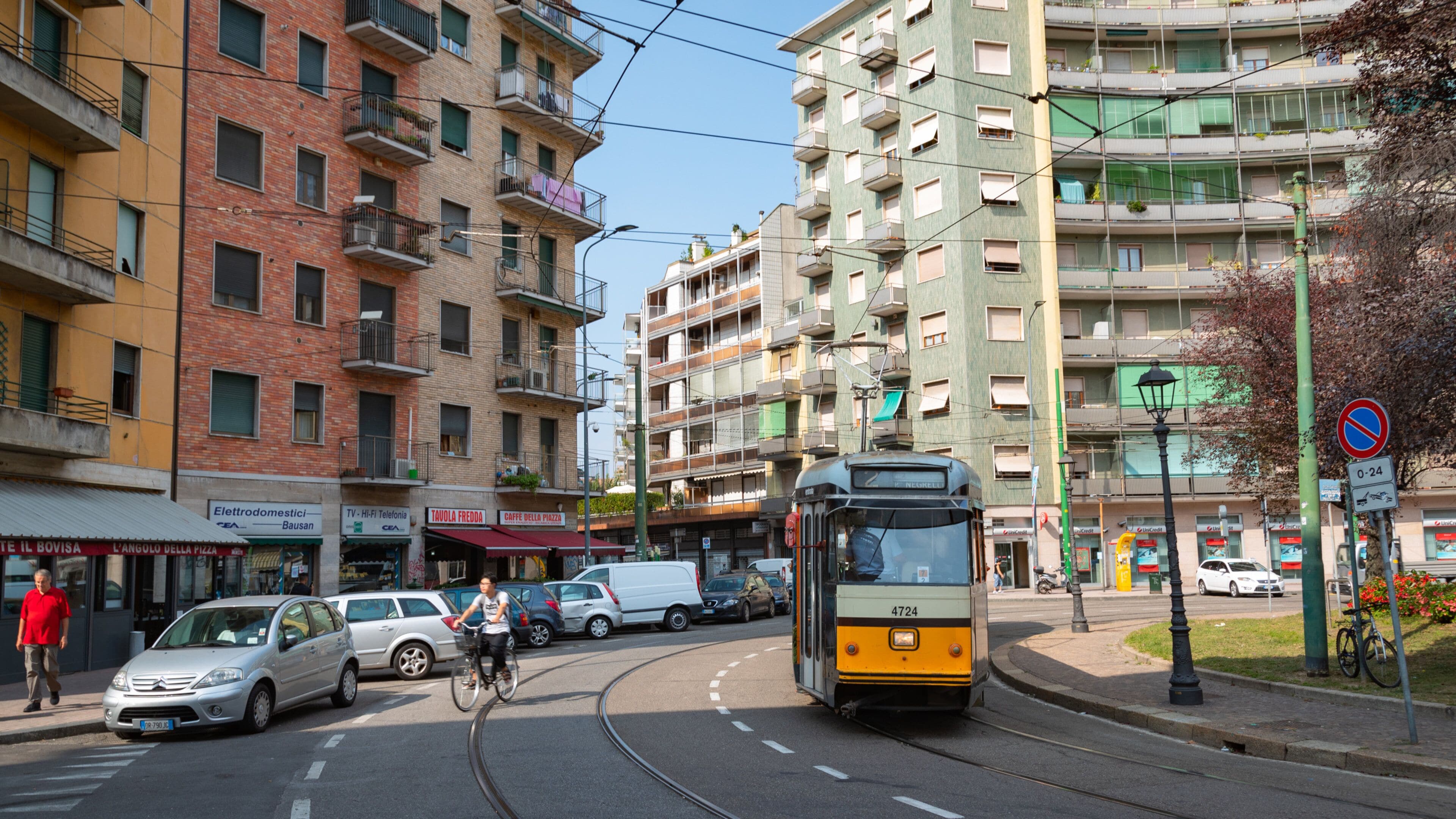 A tram on a city street with multi-story buildings, parked cars, and a no parking sign.