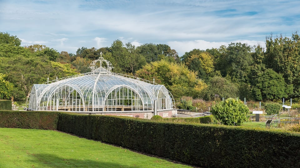 A large glass greenhouse structure surrounded by a well-maintained hedge and trees.