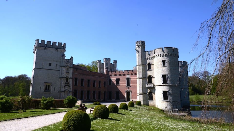 A historic castle with a moat, surrounded by well-manicured gardens and a clear blue sky.