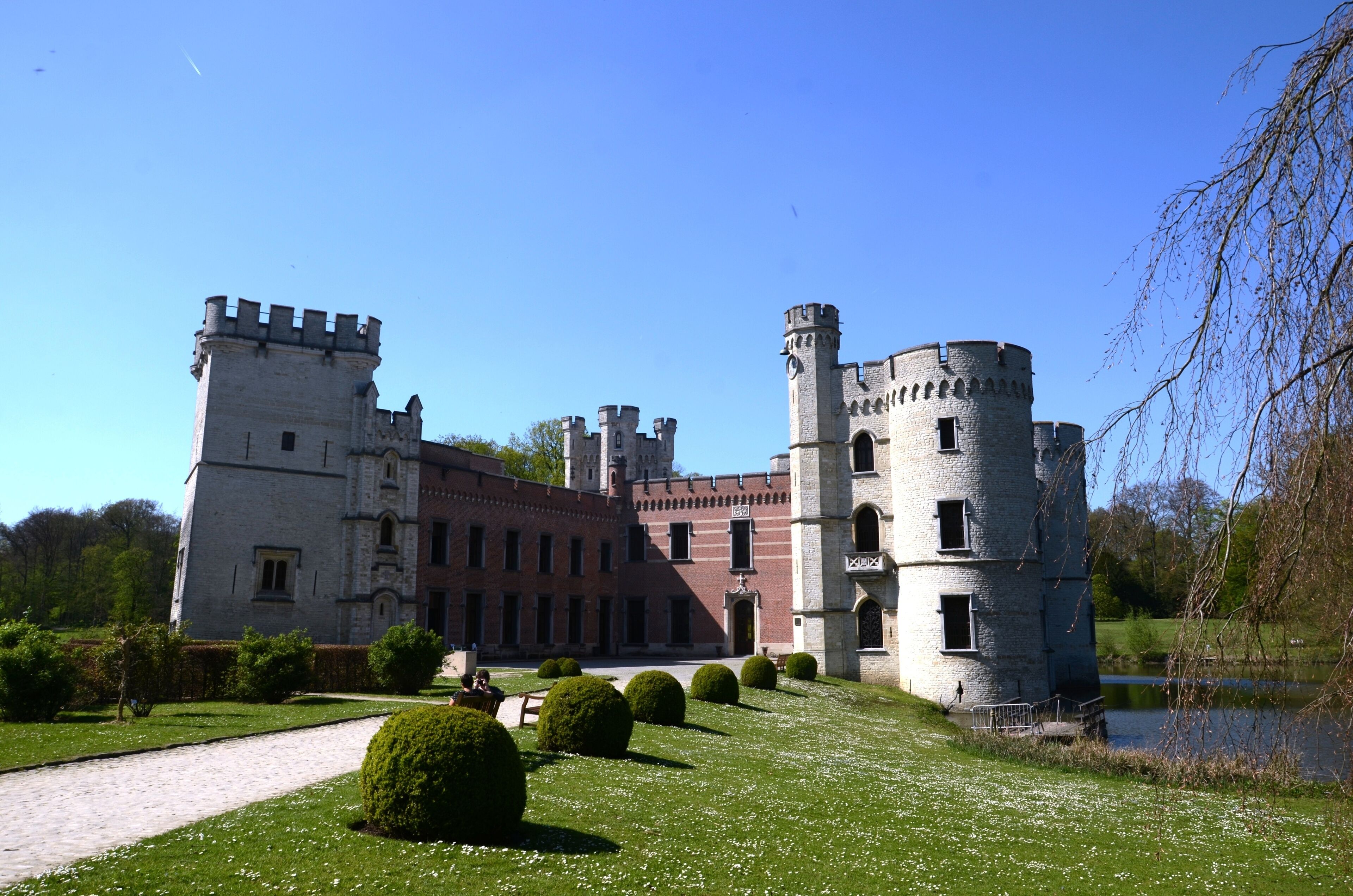 A historic castle with a moat, surrounded by well-manicured gardens and a clear blue sky.