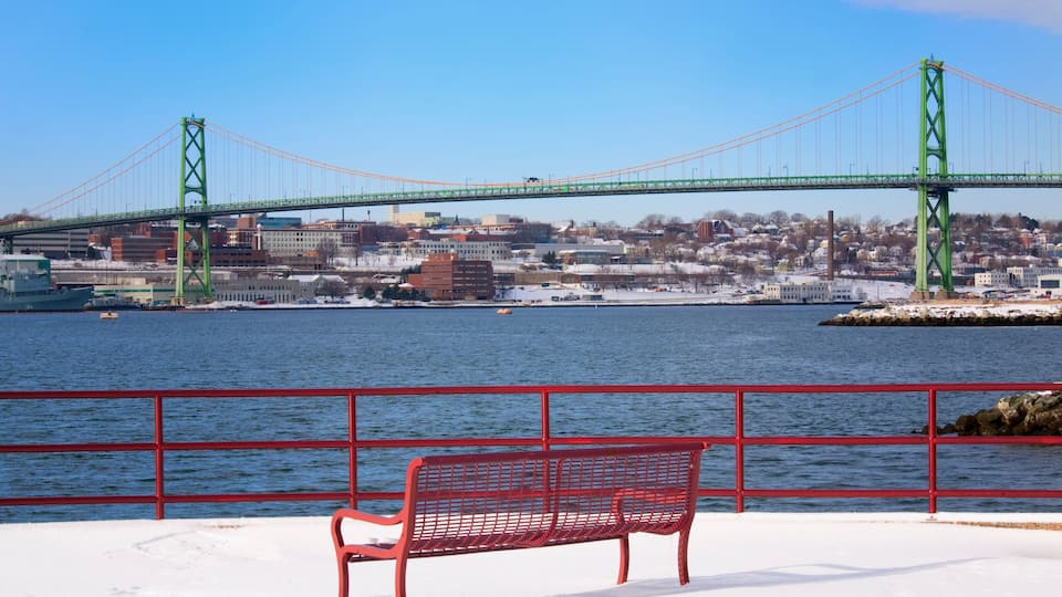A red bench by a waterfront with a green bridge in the background.