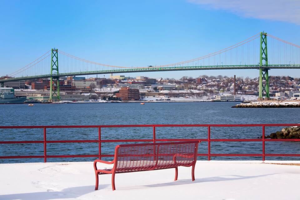 A red bench by a waterfront with a green bridge in the background.