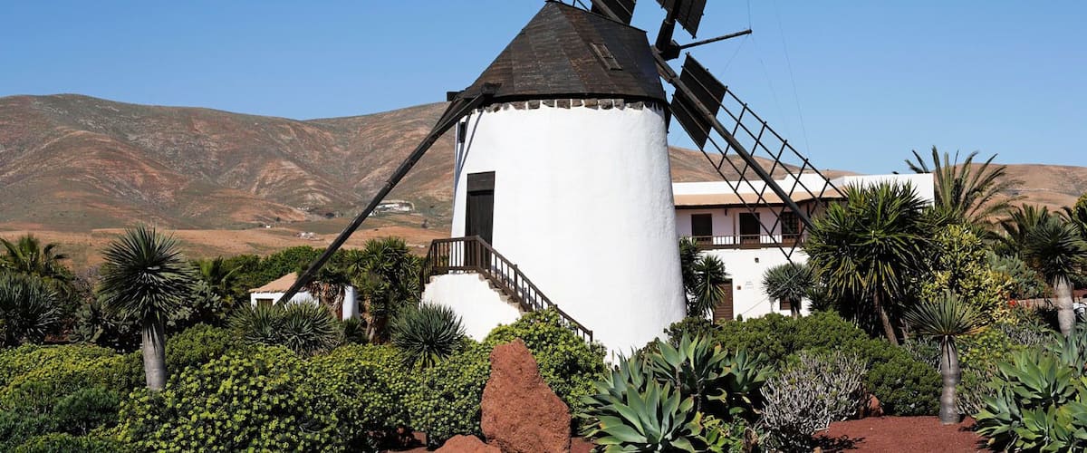 A windmill with a white body and black blades, surrounded by lush greenery and red rocks, with a mountainous backdrop.