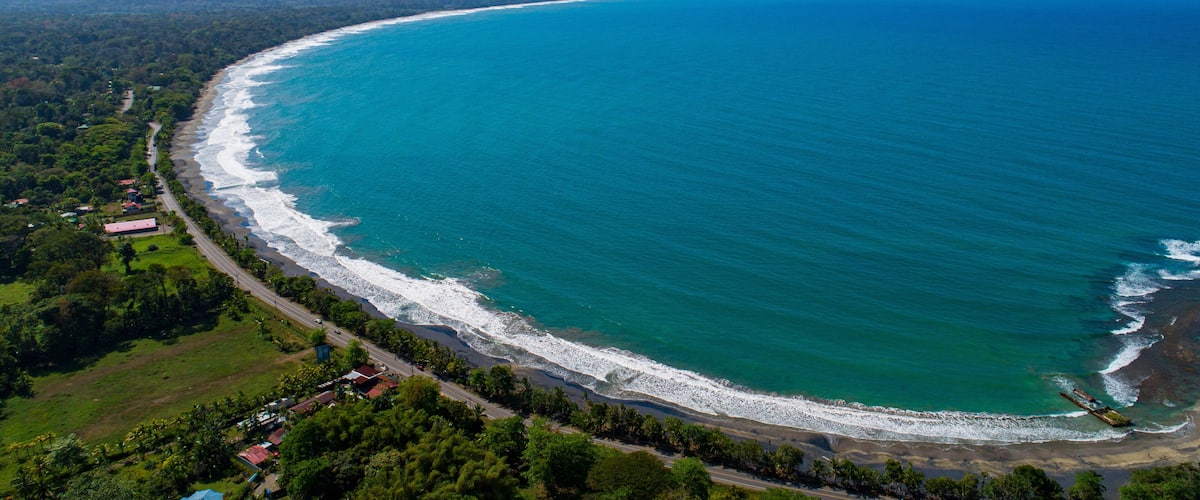 A coastal landscape with a curved shoreline, a road along the coast, and a backdrop of hills.