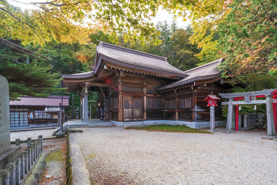 A traditional Japanese building with a wooden structure and tiled roof, surrounded by lush greenery and autumn leaves.