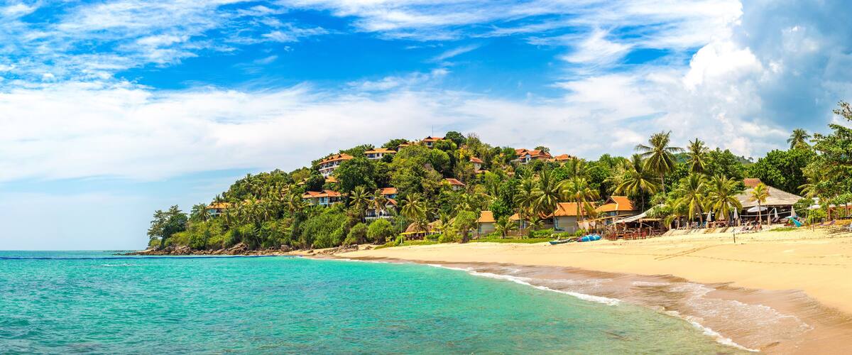 A tropical beach with turquoise waters, a sandy shore, and a hilltop with houses and palm trees.