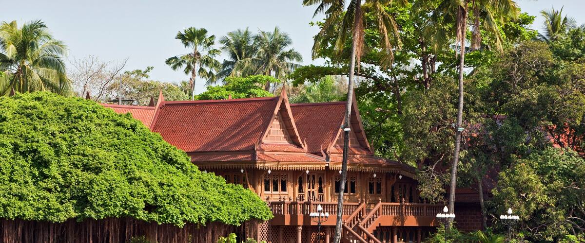 A traditional wooden building with a red roof, surrounded by lush greenery and palm trees.