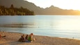 A lakeside scene with people relaxing on the shore, boats on the water, and mountains in the background.