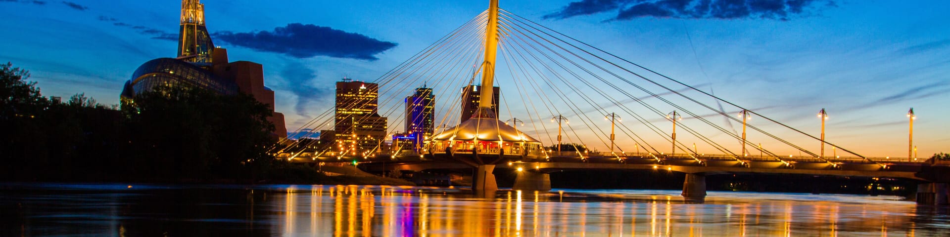 A cable-stayed bridge at dusk with city lights reflecting on a calm river.