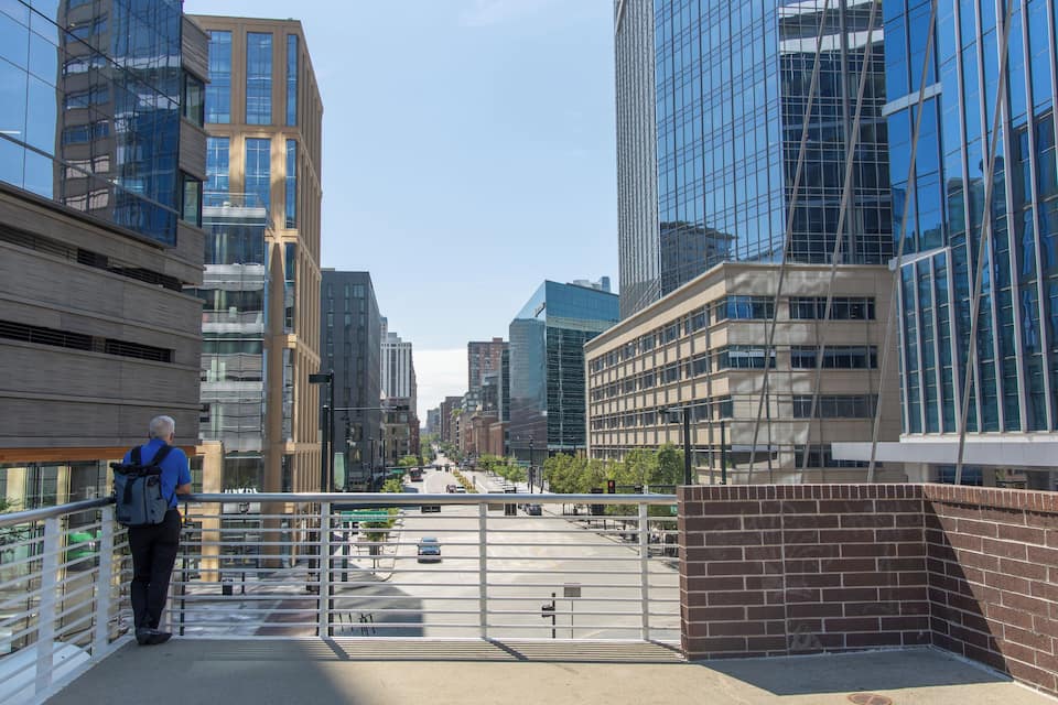 Un puente peatonal con un hombre de pie en la barandilla, observando un paisaje urbano con edificios modernos y un cielo despejado.