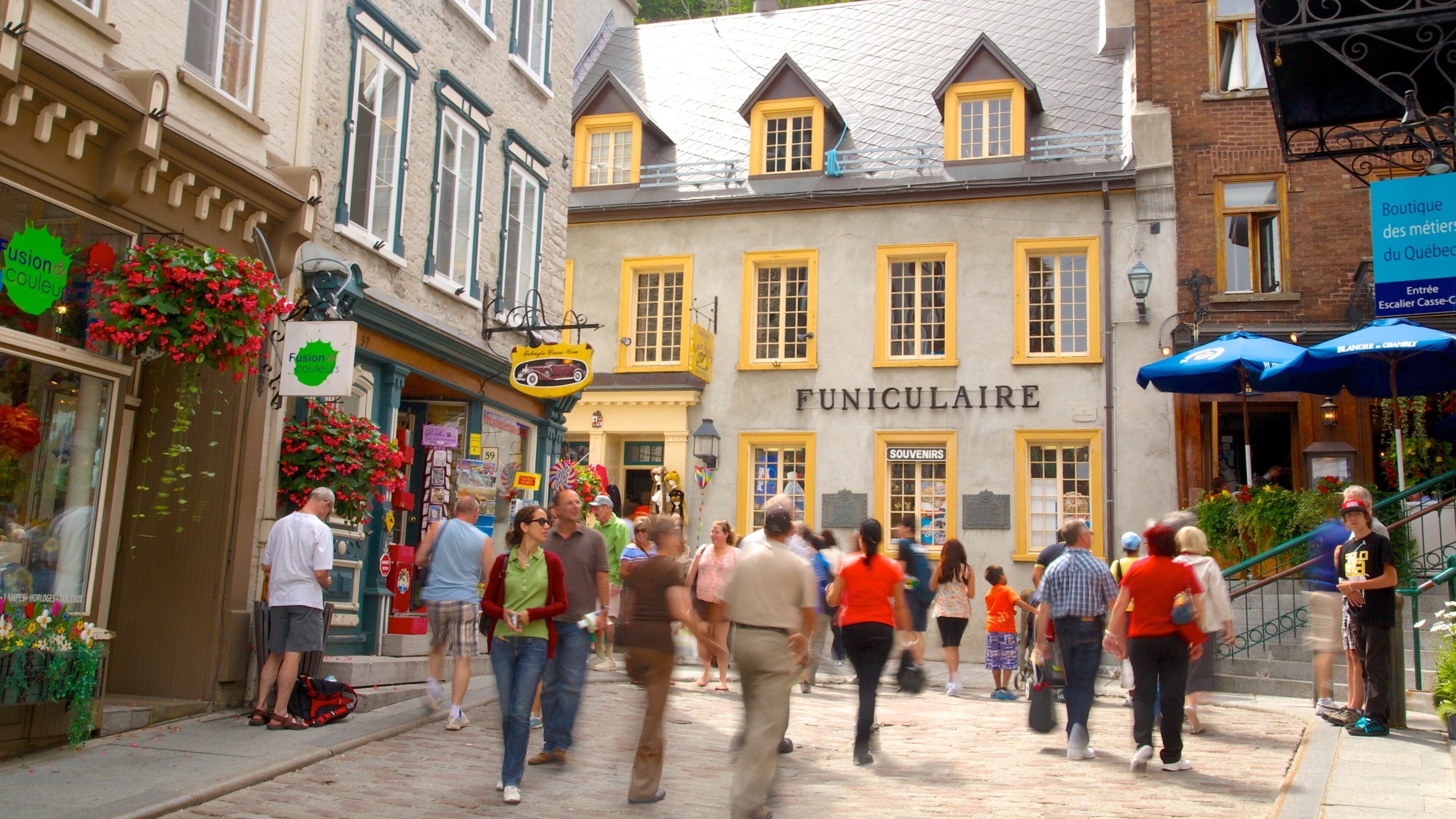 Una strada animata con persone che camminano, un edificio con la scritta "FUNICOLARE" e negozi con insegne colorate.