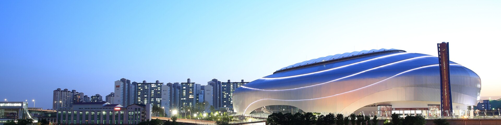 Un estadio moderno con un diseño curvo único, rodeado de edificios altos y un río tranquilo.
