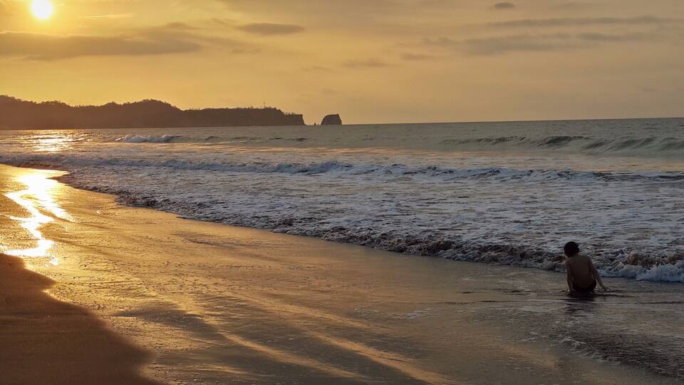 A beach with waves at sunset, a child playing in the water, and a distant shoreline with cliffs.