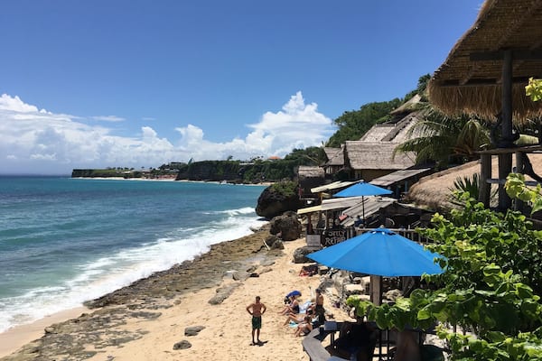 Une plage à l’eau d’un bleu limpide, avec un rivage rocheux et des constructions aux toits de chaume.