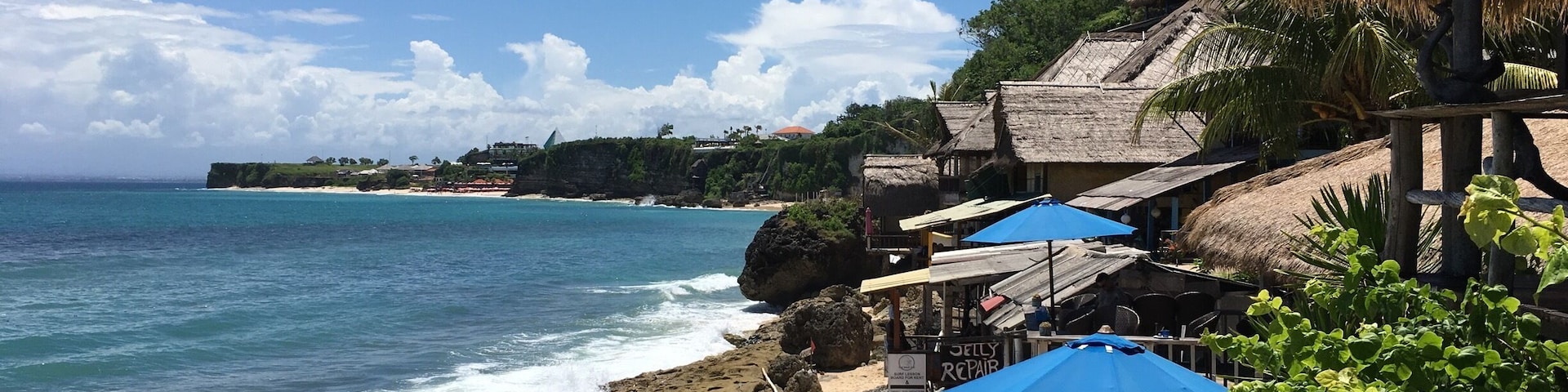 Una playa con agua azul cristalina, costa rocosa y construcciones con techo de paja.