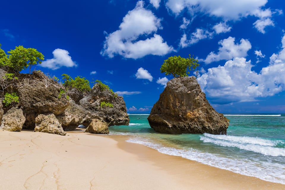 A beach with clear turquoise water, sandy shore, and large rock formations with trees on top.