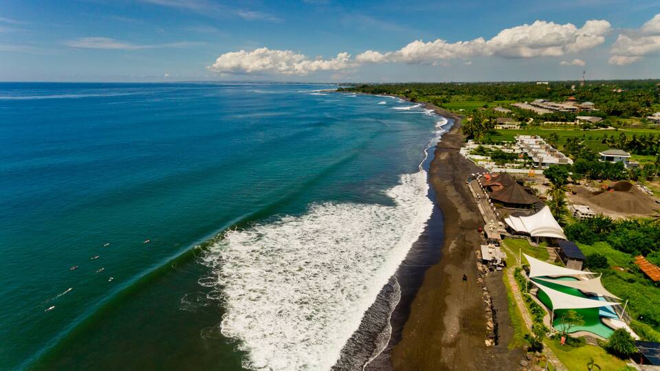 A coastal area with a beach, ocean waves, and a resort with buildings and a pool.