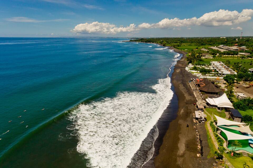 A coastal area with a beach, ocean waves, and a resort with buildings and a pool.