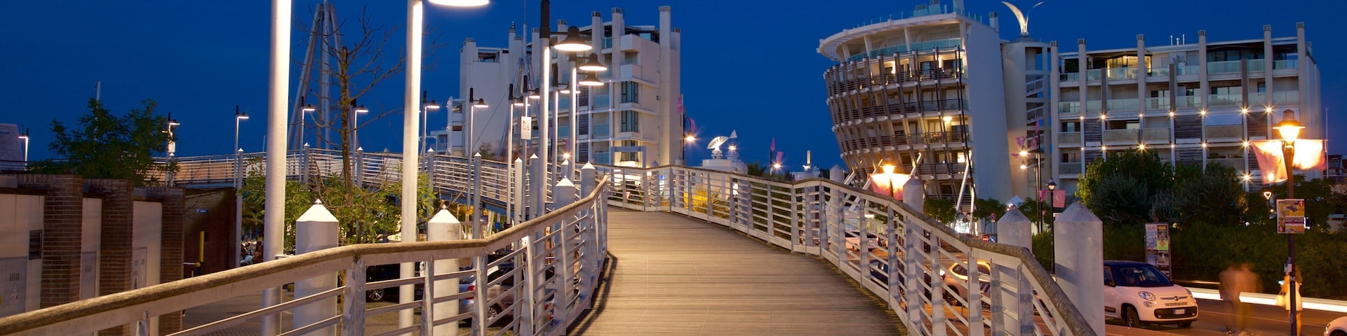 A wooden bridge at dusk with street lamps and modern buildings in the background.