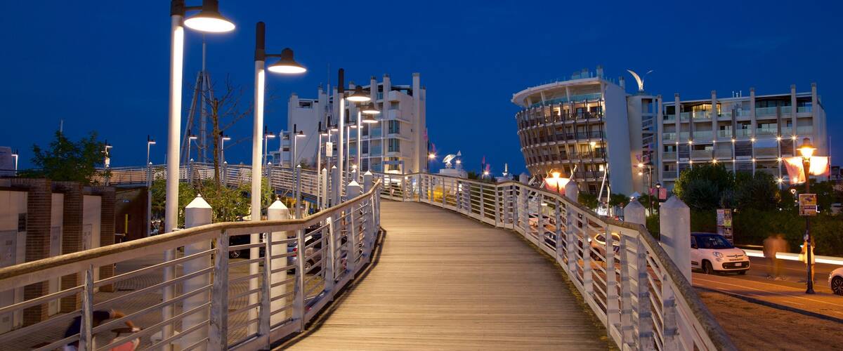 A wooden bridge at dusk with street lamps and modern buildings in the background.