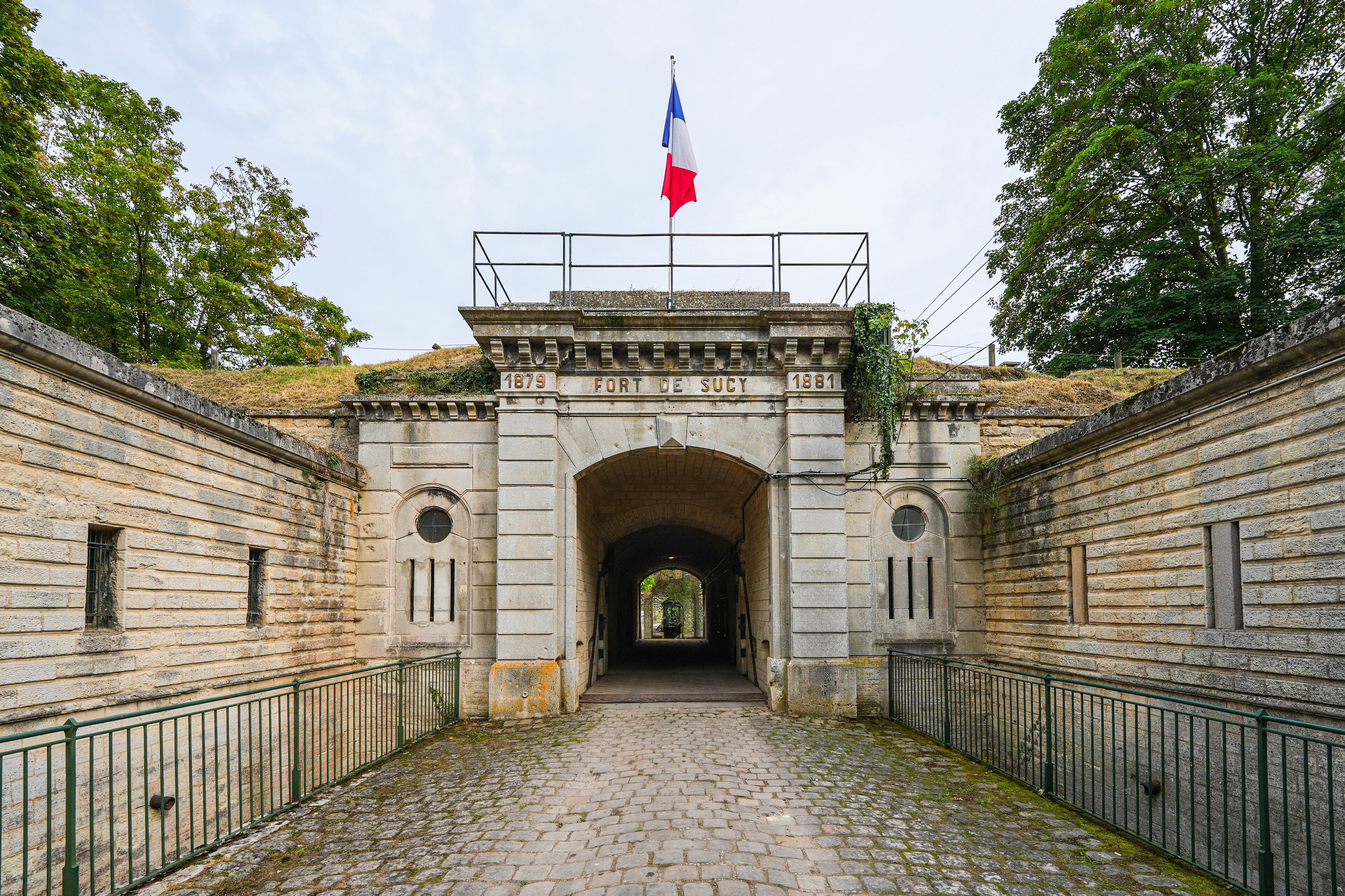 An entrance to Fort de Sucy with a French flag, stone walls, and cobblestone path.