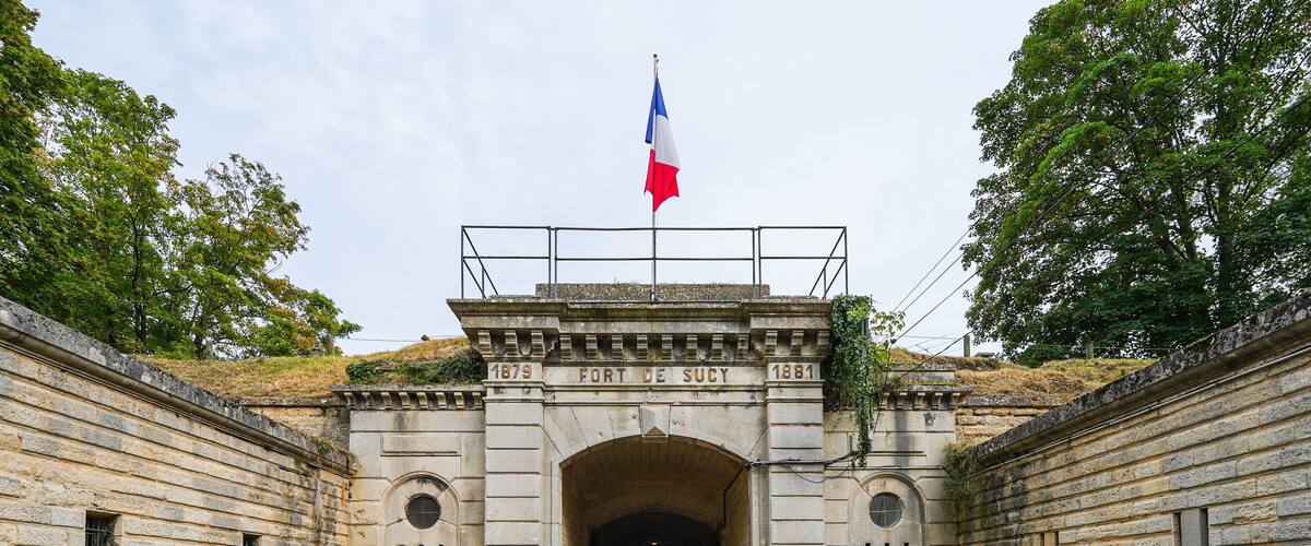 An entrance to Fort de Sucy with a French flag, stone walls, and cobblestone path.