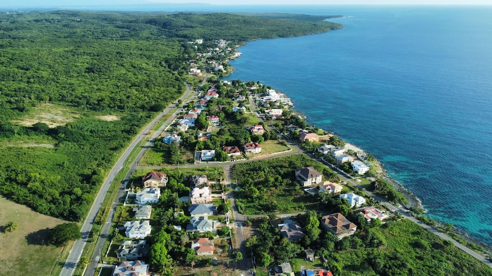 A coastal residential area with houses along the shoreline, surrounded by dense greenery and a clear blue sea.