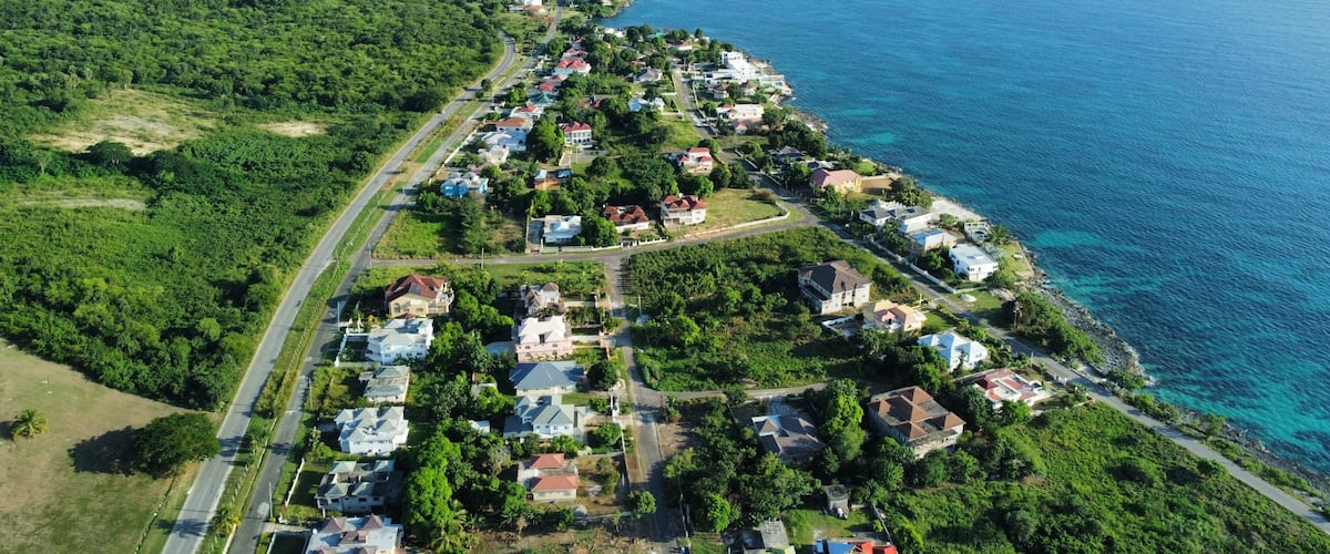 A coastal residential area with houses along the shoreline, surrounded by dense greenery and a clear blue sea.